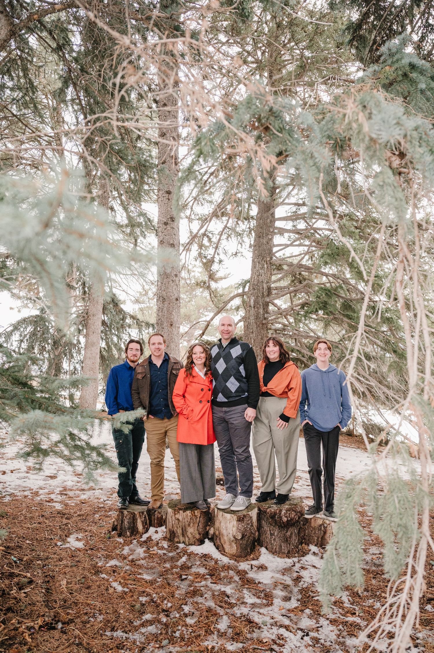 A group of people standing together on tree stumps in a snow-dusted forest wearing winter outdoor clothing.