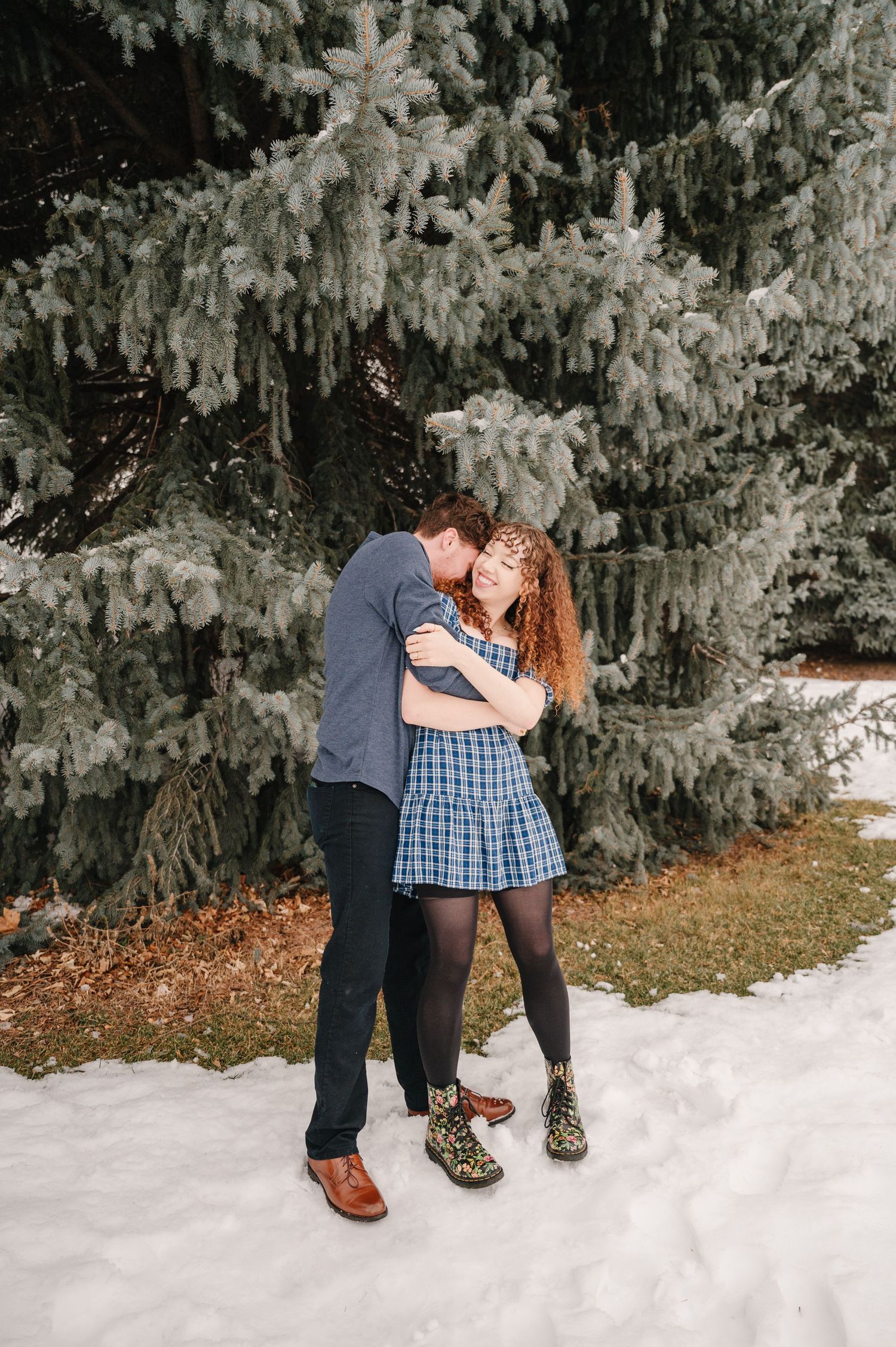 A romantic winter scene shows a couple embracing under snow-covered pine trees in a natural outdoor setting.
