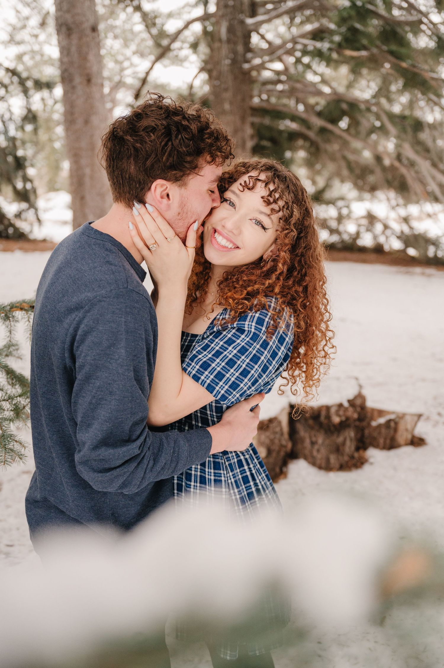 Romantic winter scene in a snowy forest with a couple sharing an intimate moment in their blue plaid and navy outfits.