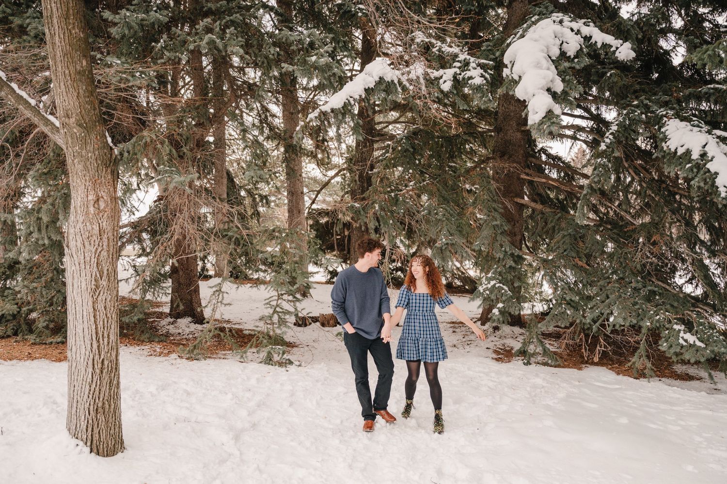 A couple walks together through a snowy forest path with evergreen trees surrounding them in winter.