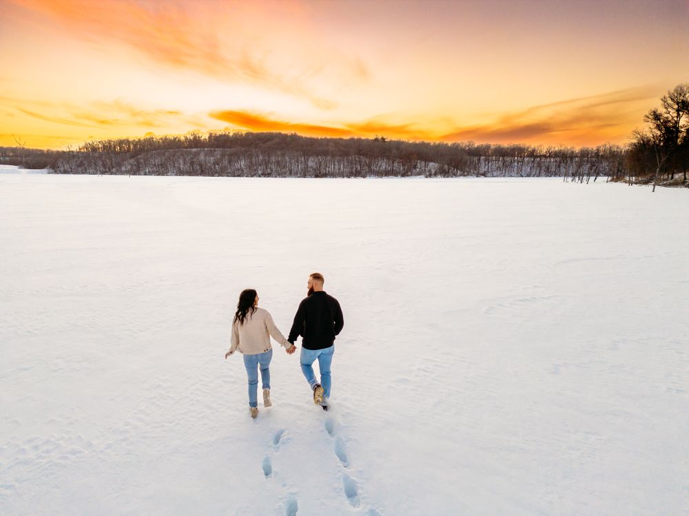 Couple walking through snowy landscape with vibrant orange sunset sky.