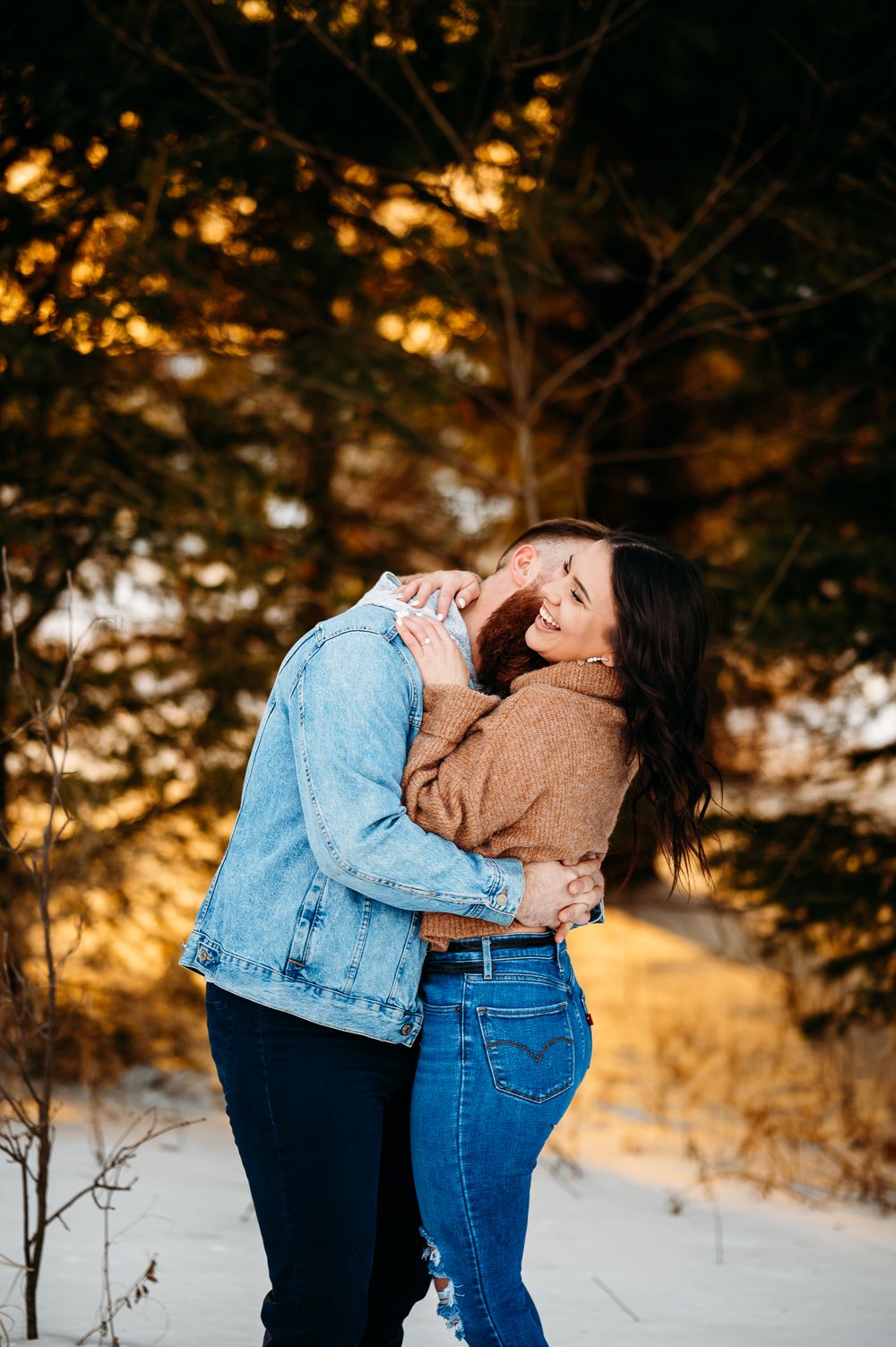 A romantic couple embracing and laughing in a snowy winter forest setting during golden hour.