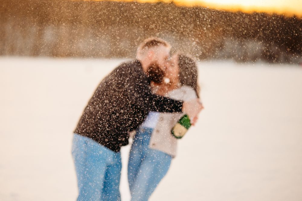 Joyful couple celebrating with champagne spray in the snow at sunset.