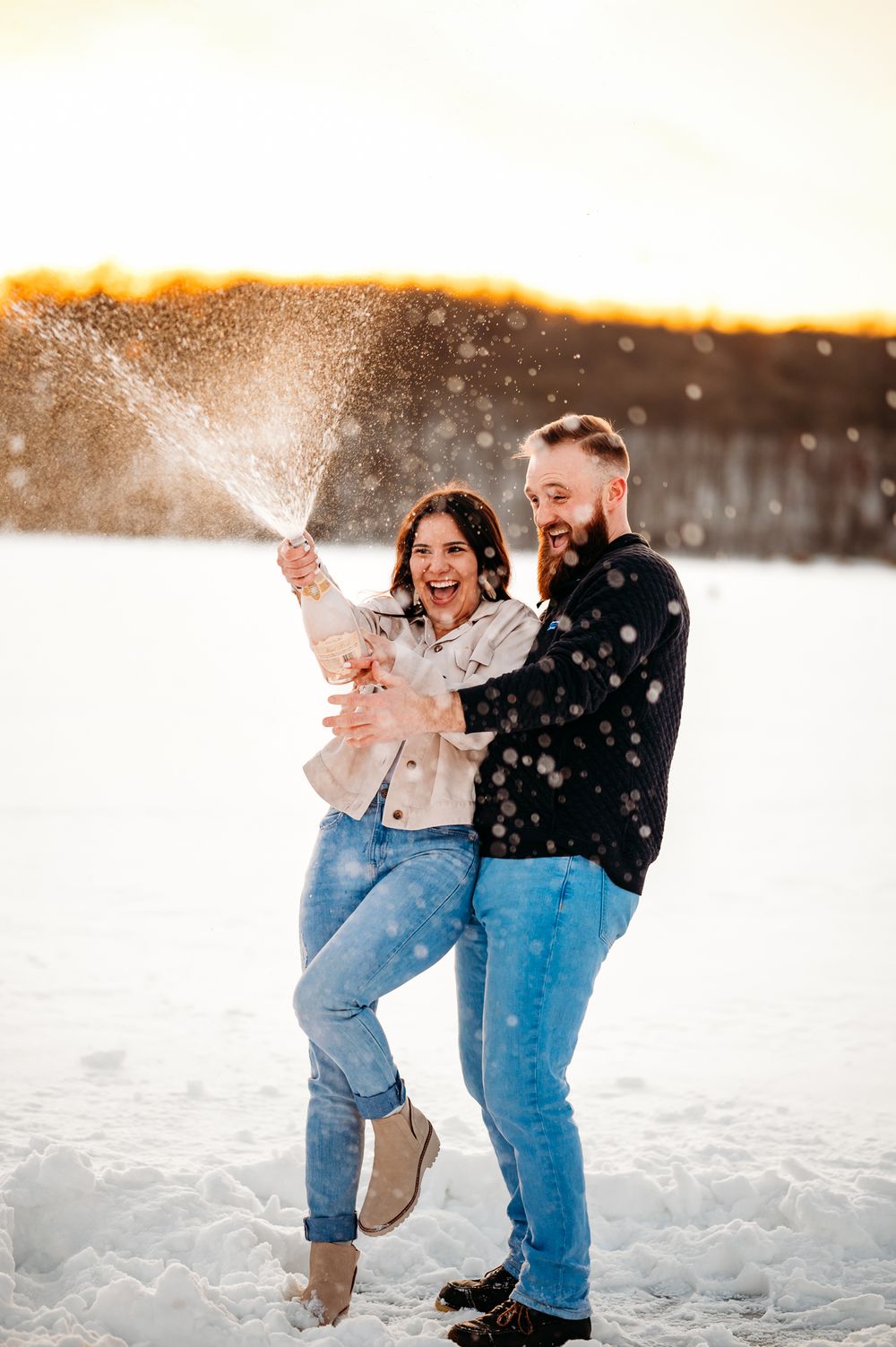 Happy couple popping champagne and celebrating in snowy winter landscape.