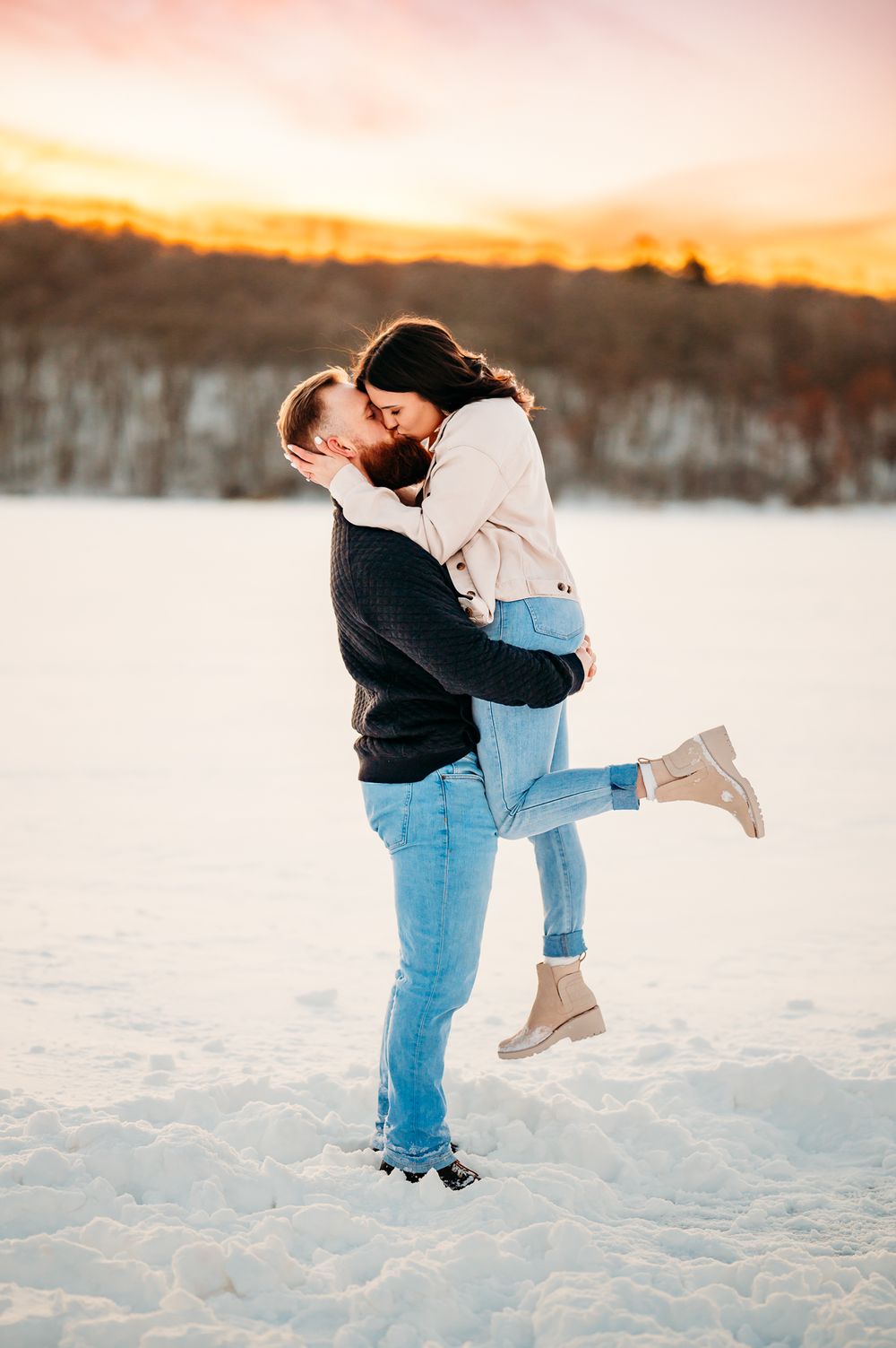 Couple sharing a romantic lift and kiss against a winter sunset backdrop.