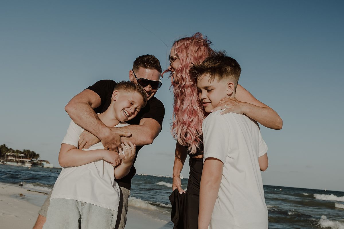 Family of four laughing and hugging on a sunny beach, with a pink-haired woman and two young boys.