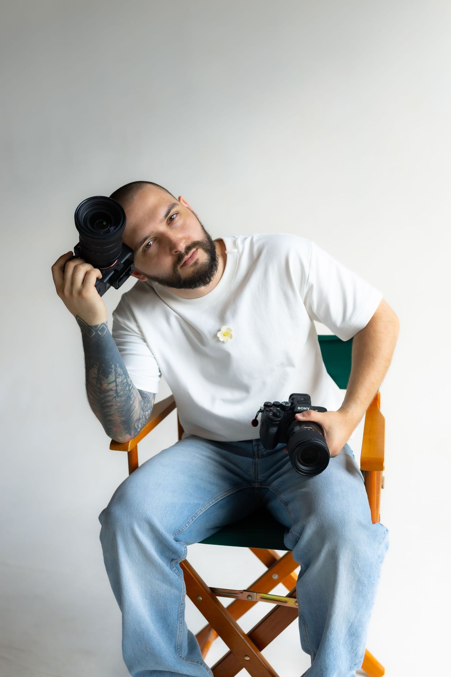 Professional photographer in white shirt and jeans holding two cameras while sitting on a wooden director's chair.