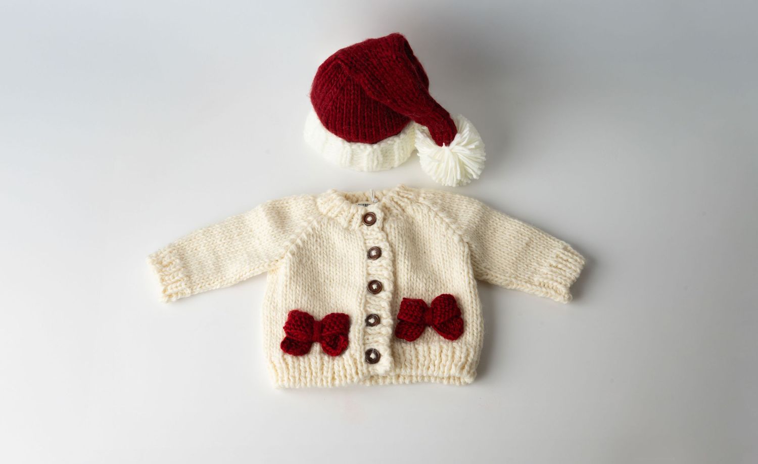 Handknit baby cardigan in cream white with red bows and matching red knit hat for winter wear.