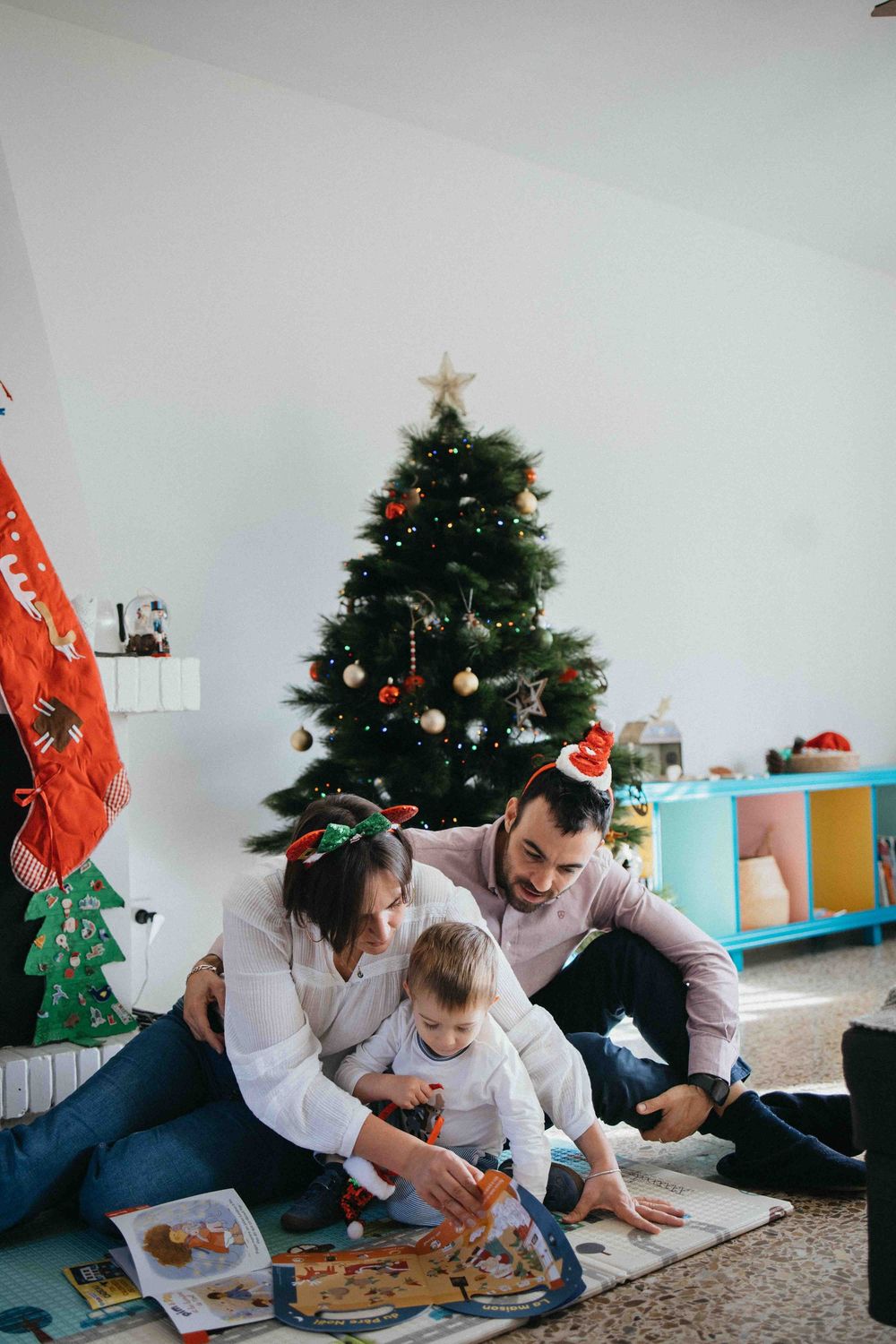 Parent and child in festive Santa hats sit together on floor looking at a book during holiday season.