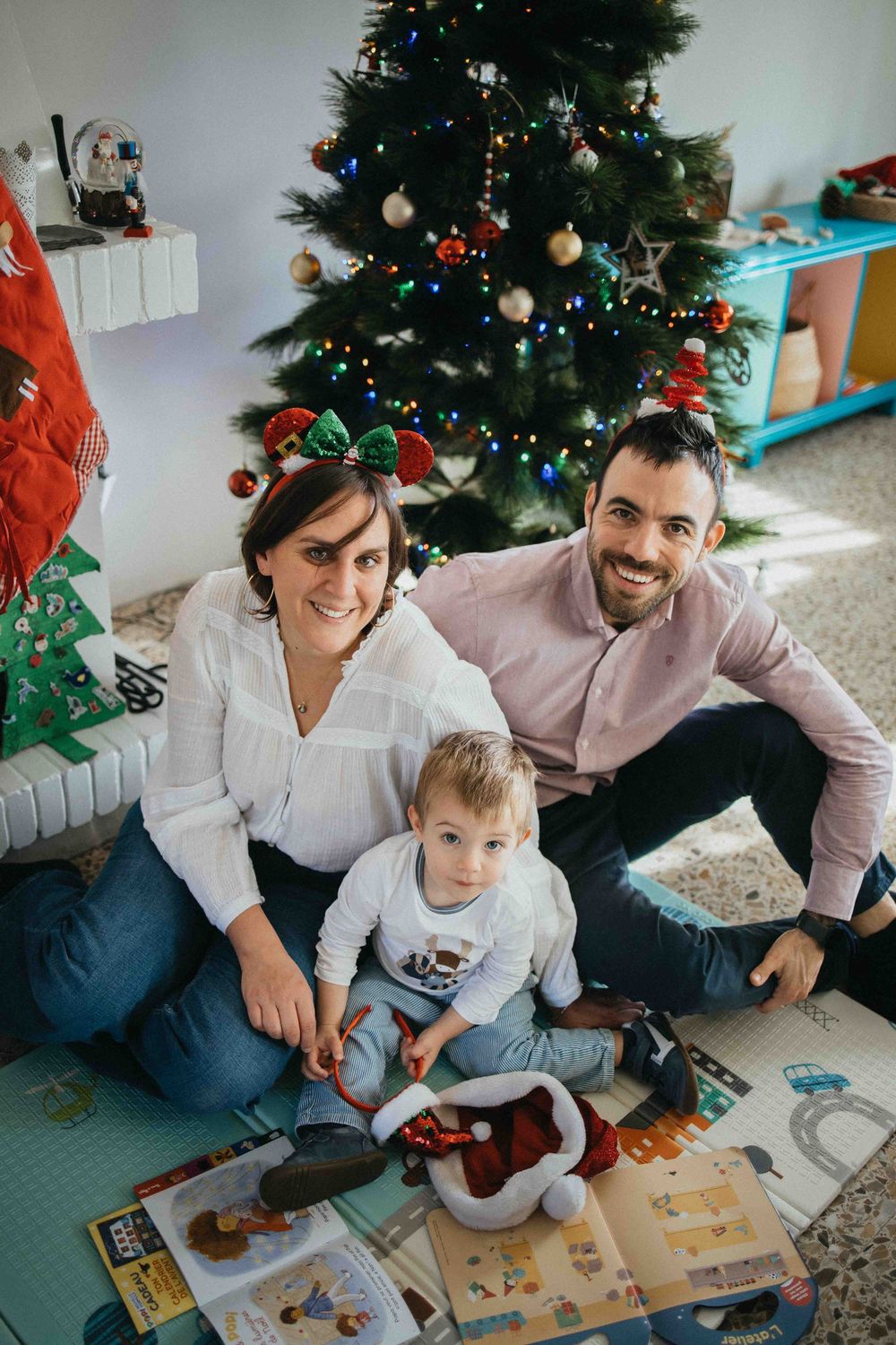 A family sits together by a decorated Christmas tree while looking at books and toys on the floor.