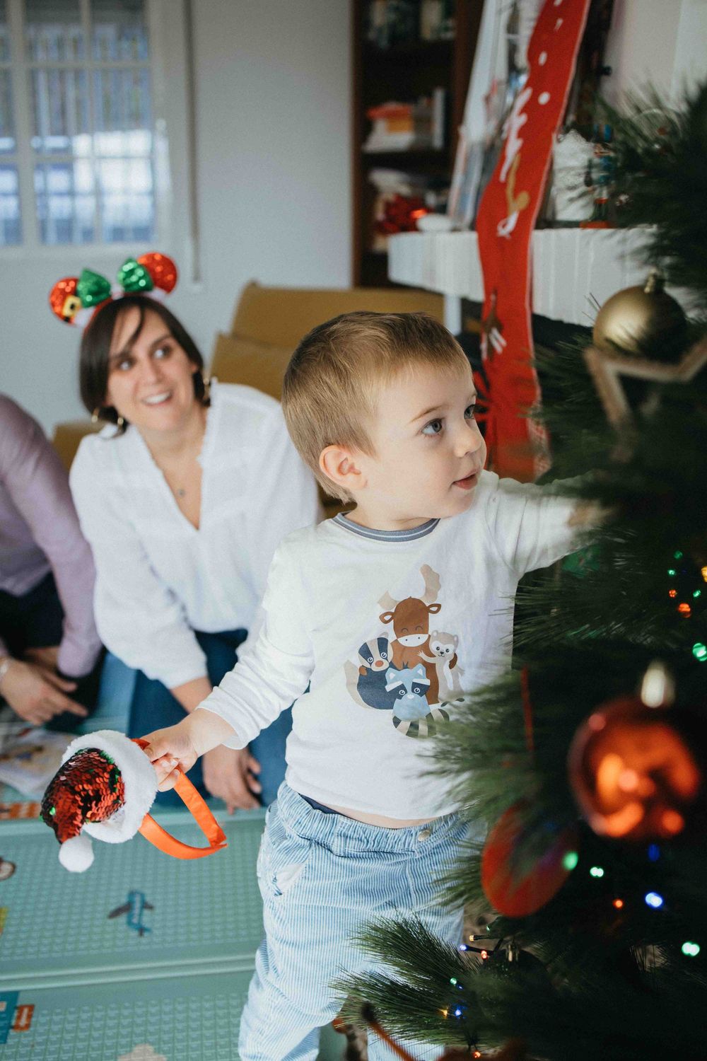 A small child in a white sweater and jeans decorates a Christmas tree with ornaments in a sequence of photos.