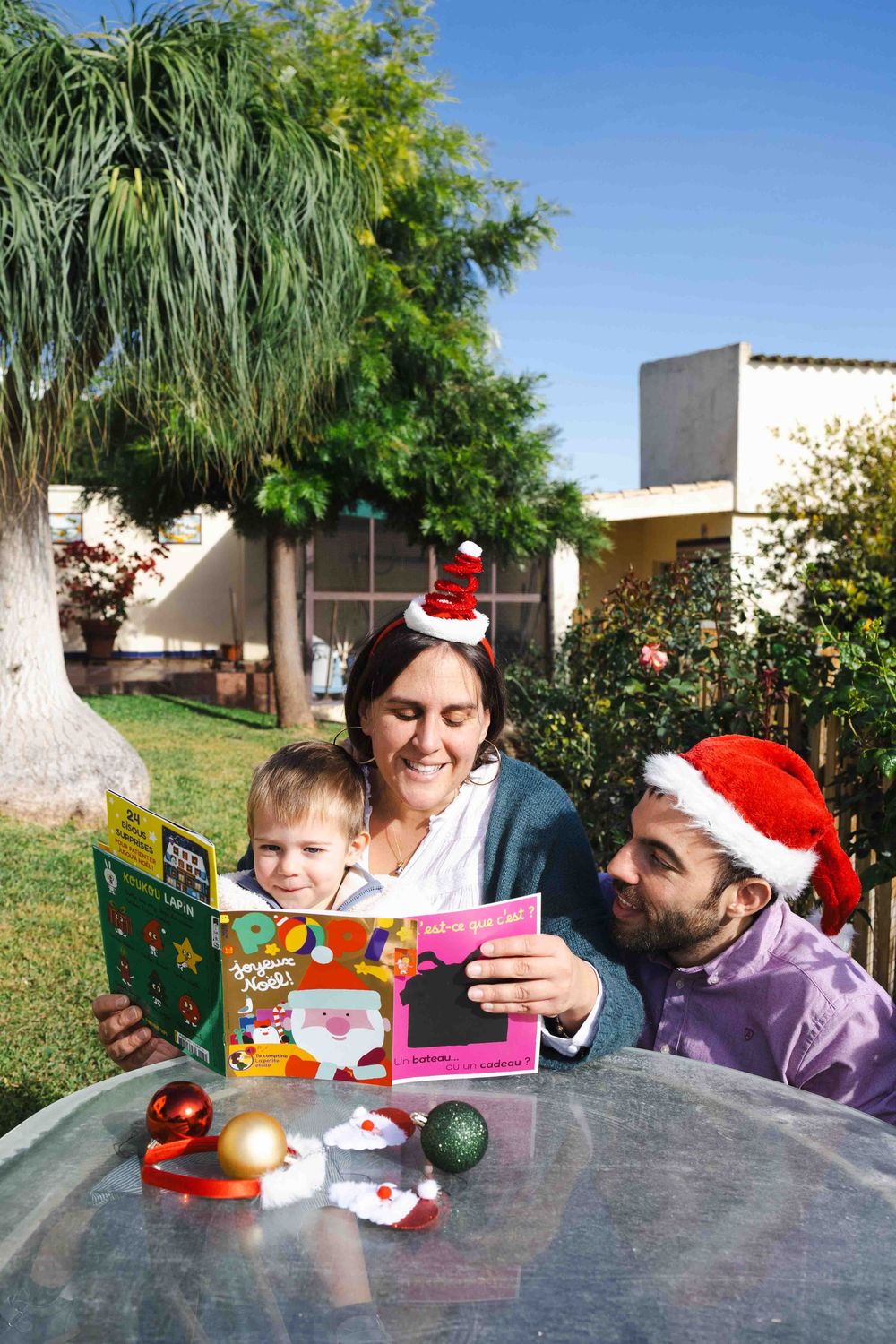 People in Santa hats gather around a table outdoors with holiday decorations and toys on a sunny day.