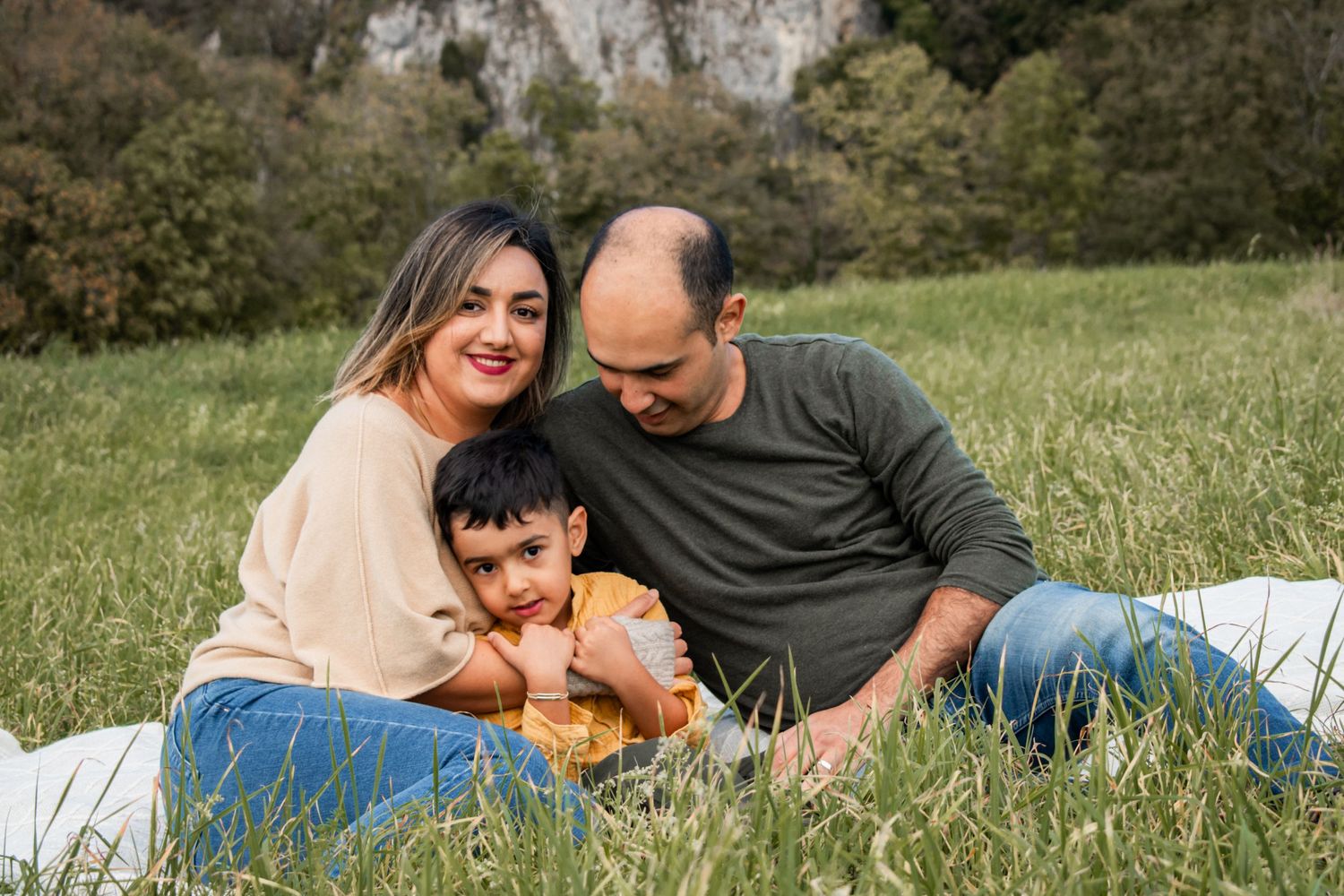 A family enjoys a relaxing moment together on a picnic blanket in a grassy field with rocky cliffs in the background.