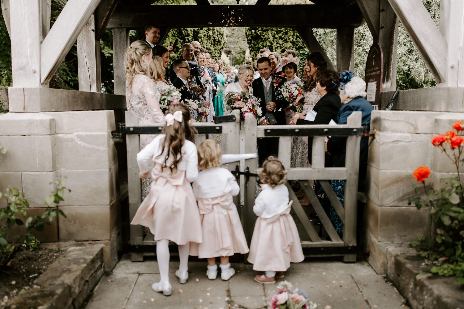 Three small children in white dresses peek through a wooden gate at a garden wedding celebration.