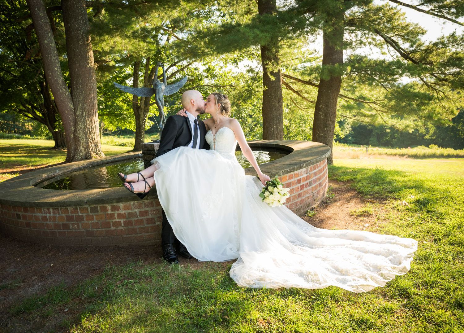 A couple in wedding attire sits together on a bench in a sunlit garden setting during their special day.
