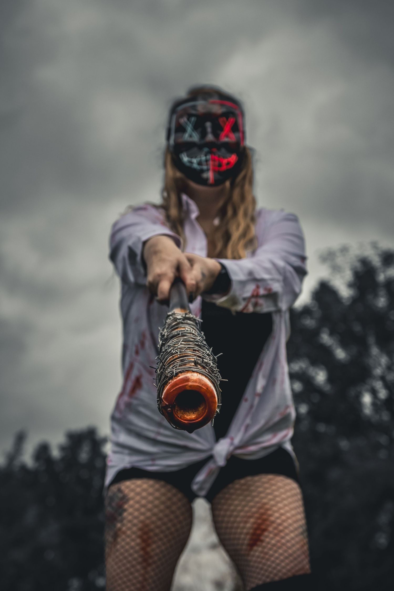 Woman in bloody Halloween costume holding barbed wire bat toward camera.