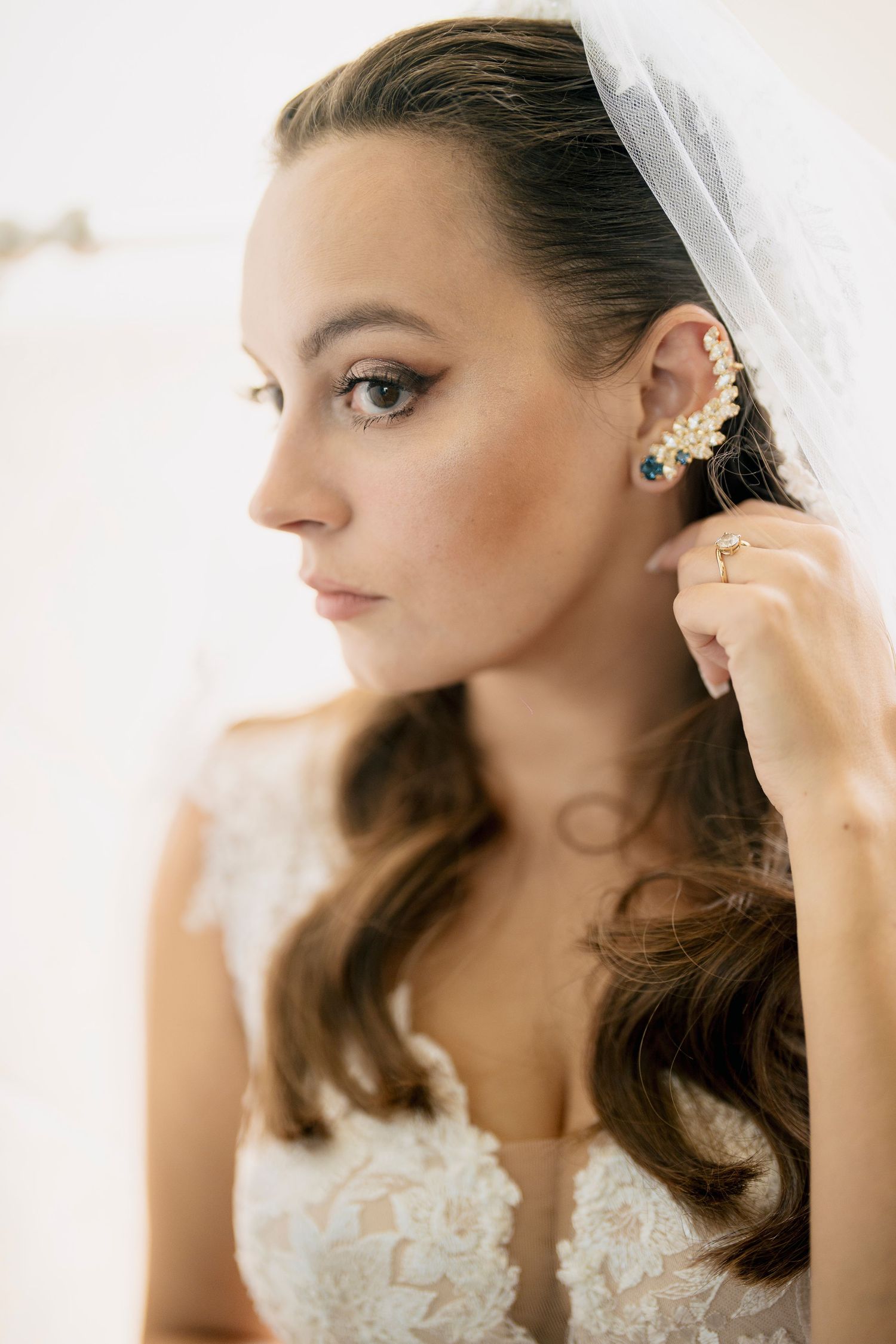 A bride in a white lace wedding dress adjusts her earrings while getting ready for her special day.