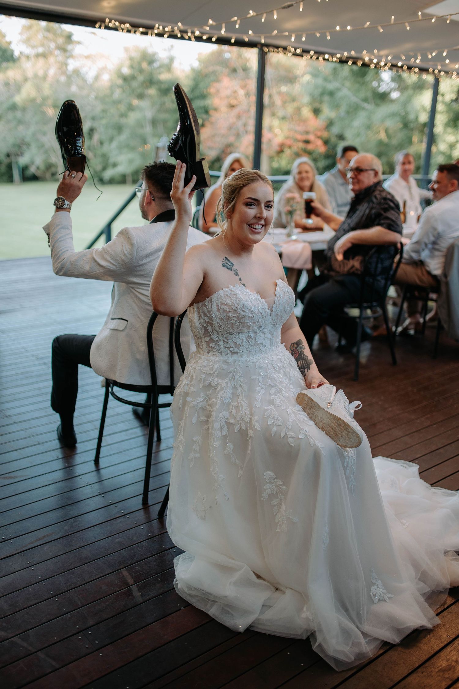 Joyful wedding reception scene with bride dancing and twirling in a beautiful white lace gown on wooden deck under lights.