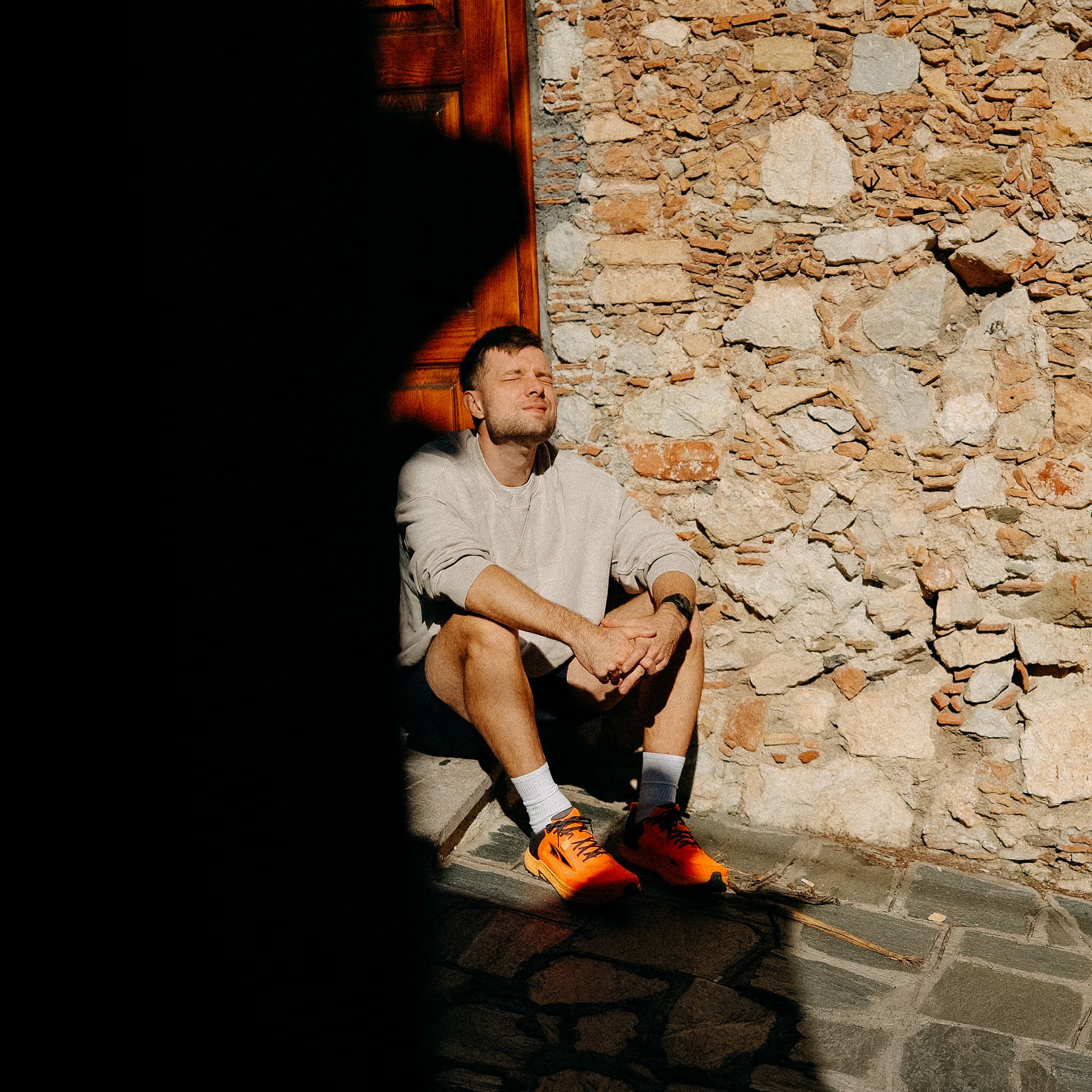 Figure sitting against rustic stone wall in dramatic lighting wearing casual clothes and colorful shoes.