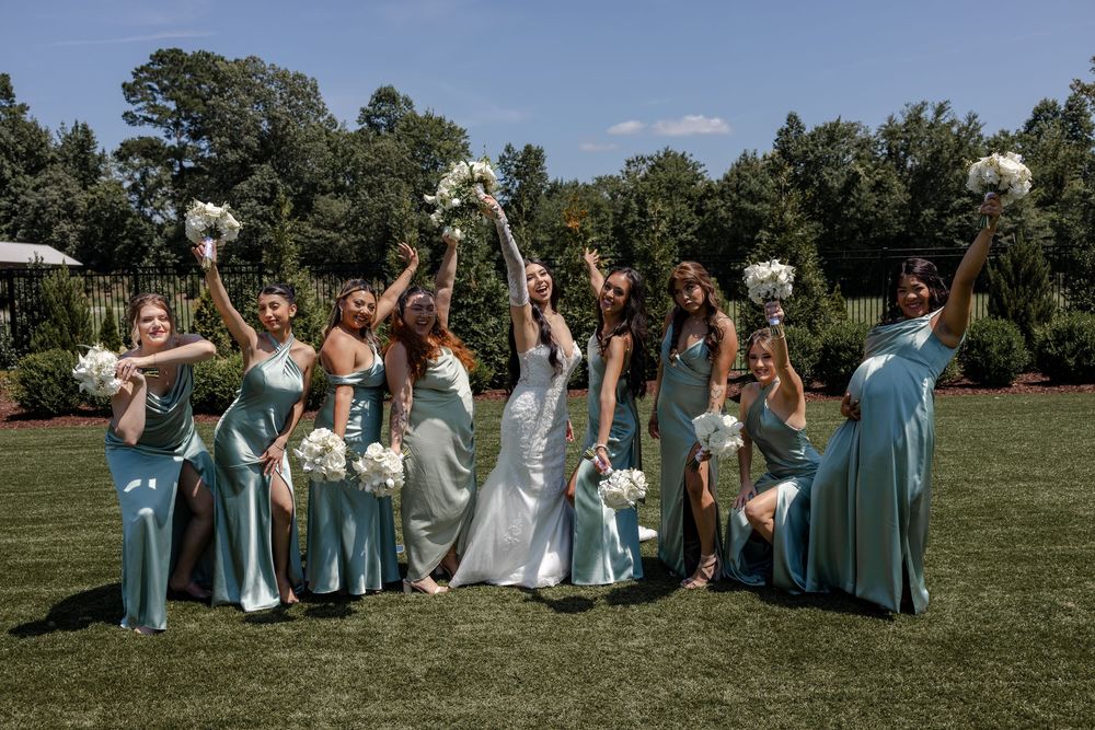 Wedding party in light blue dresses poses playfully on grassy lawn during outdoor celebration.