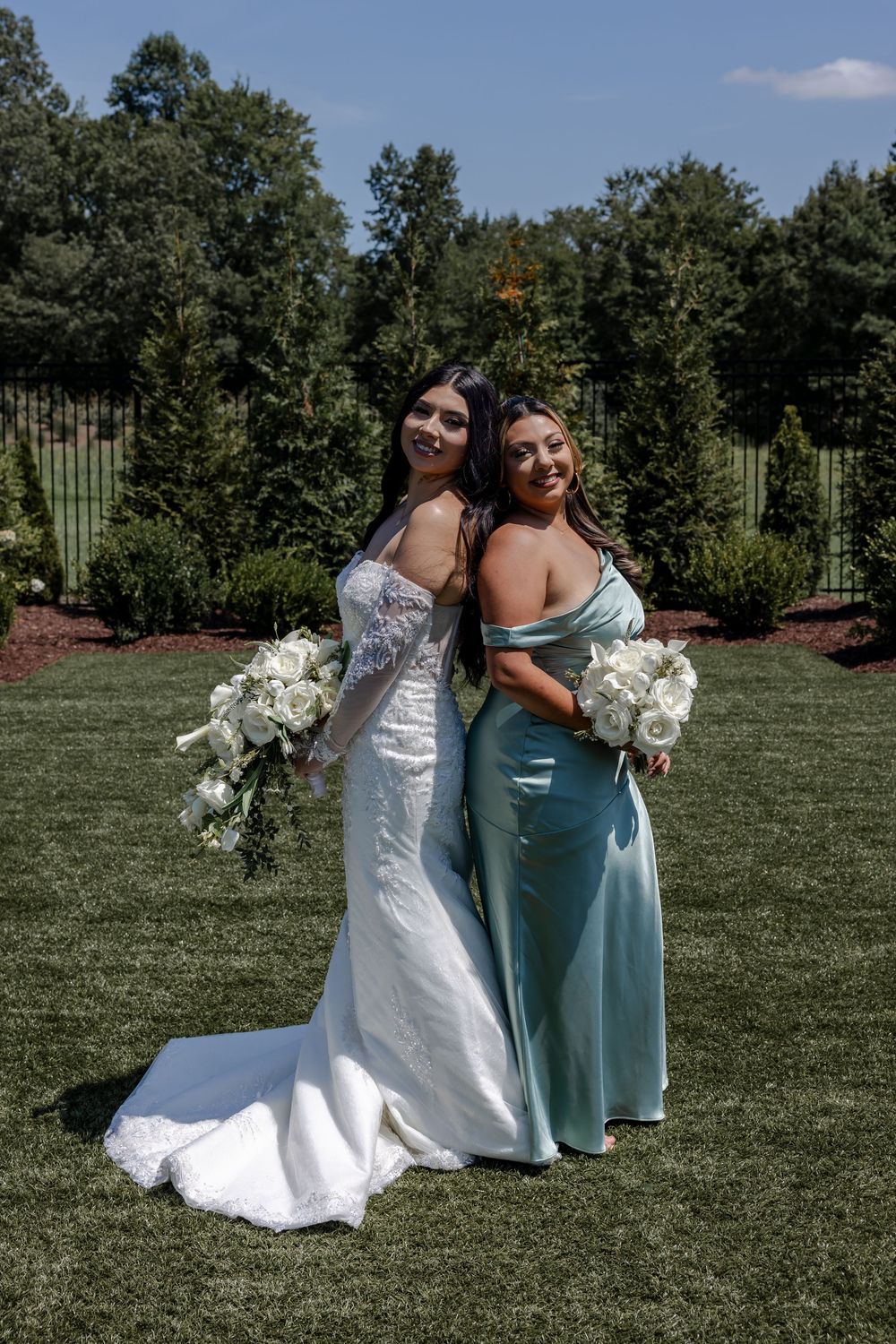 Wedding party poses on manicured lawn with white and sage green dresses holding floral bouquets.