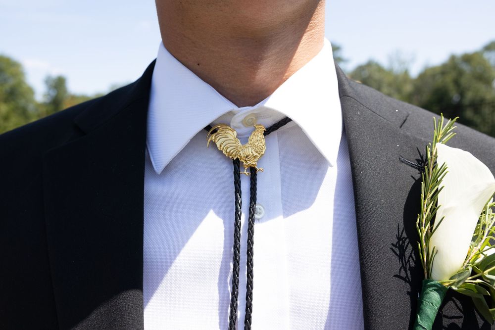 A row of groomsmen in black suits and bolo ties with white floral boutonnieres stand together at a wedding.