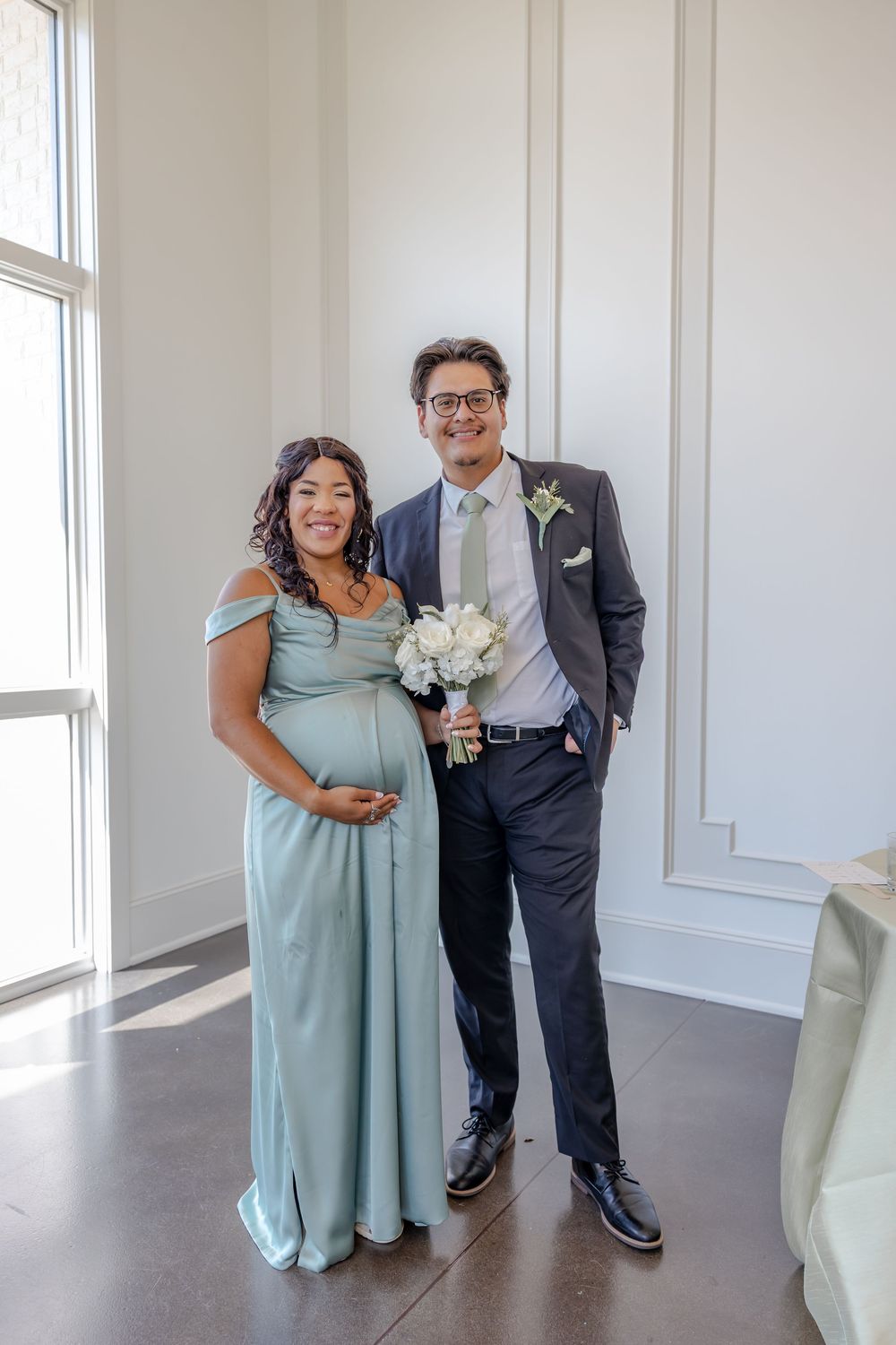 A couple poses in formal attire with a white bouquet against a bright wall with elegant molding.