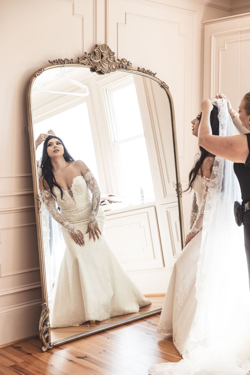 An ornate vintage mirror reflects a bride in an elegant white lace wedding gown in a light-filled dressing room.