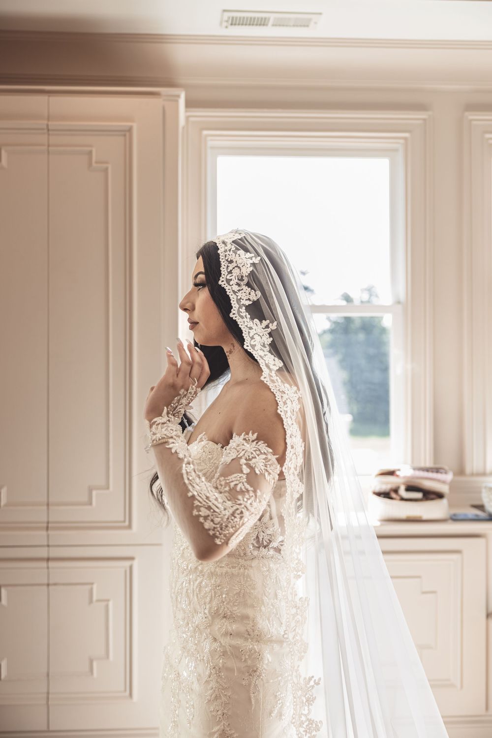 Elegant lace wedding dress and veil photographed in soft window light against white paneled walls.