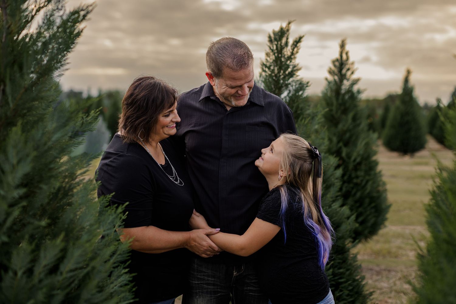 A couple embraces in front of evergreen trees during a cloudy outdoor photo session.