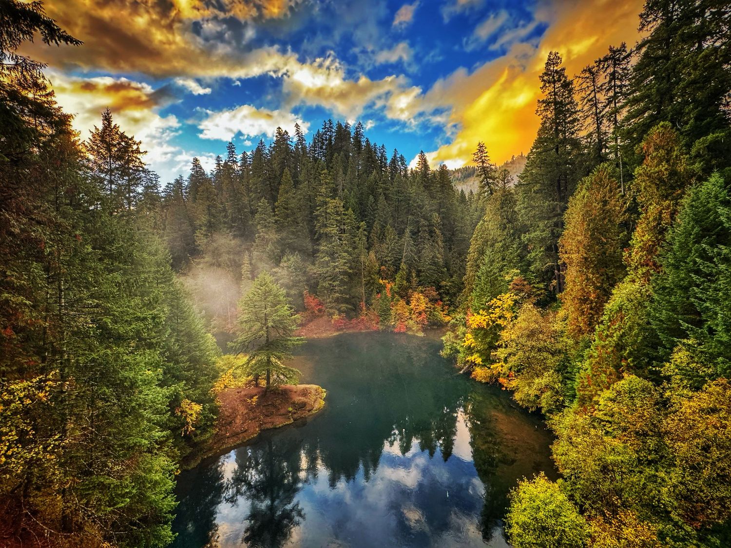 Dramatic sunset reflects on a calm lake surrounded by evergreen forest and autumn colors.