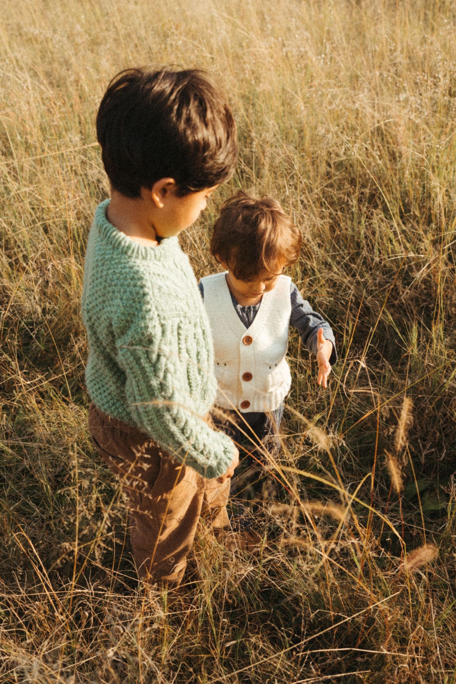 Two children in sweaters explore through dry autumn grass.
