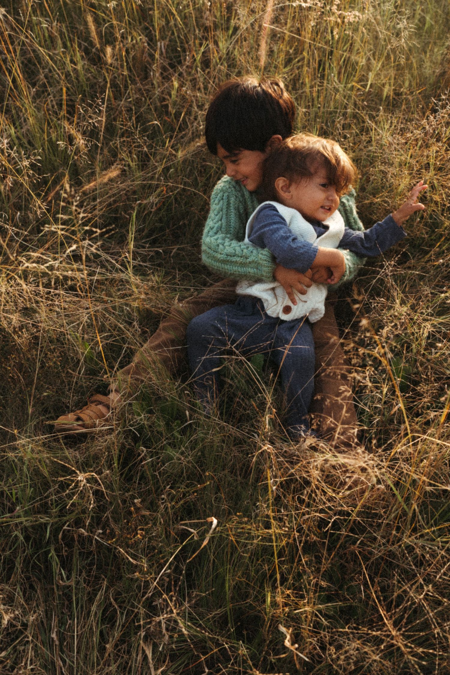 Children share a sweet moment in tall grass during golden hour.
