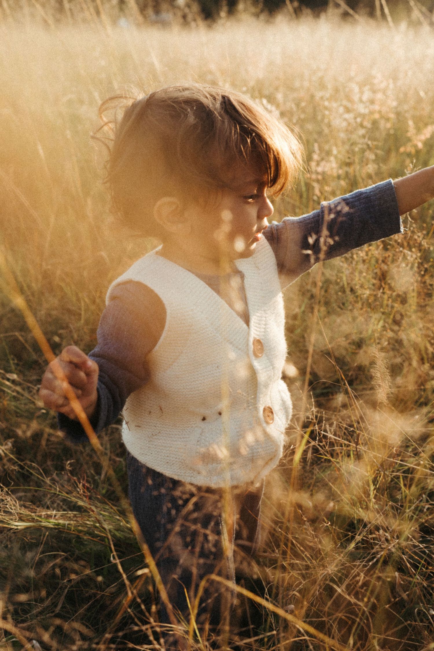Child explores tall grass wearing white button-up vest at golden hour.