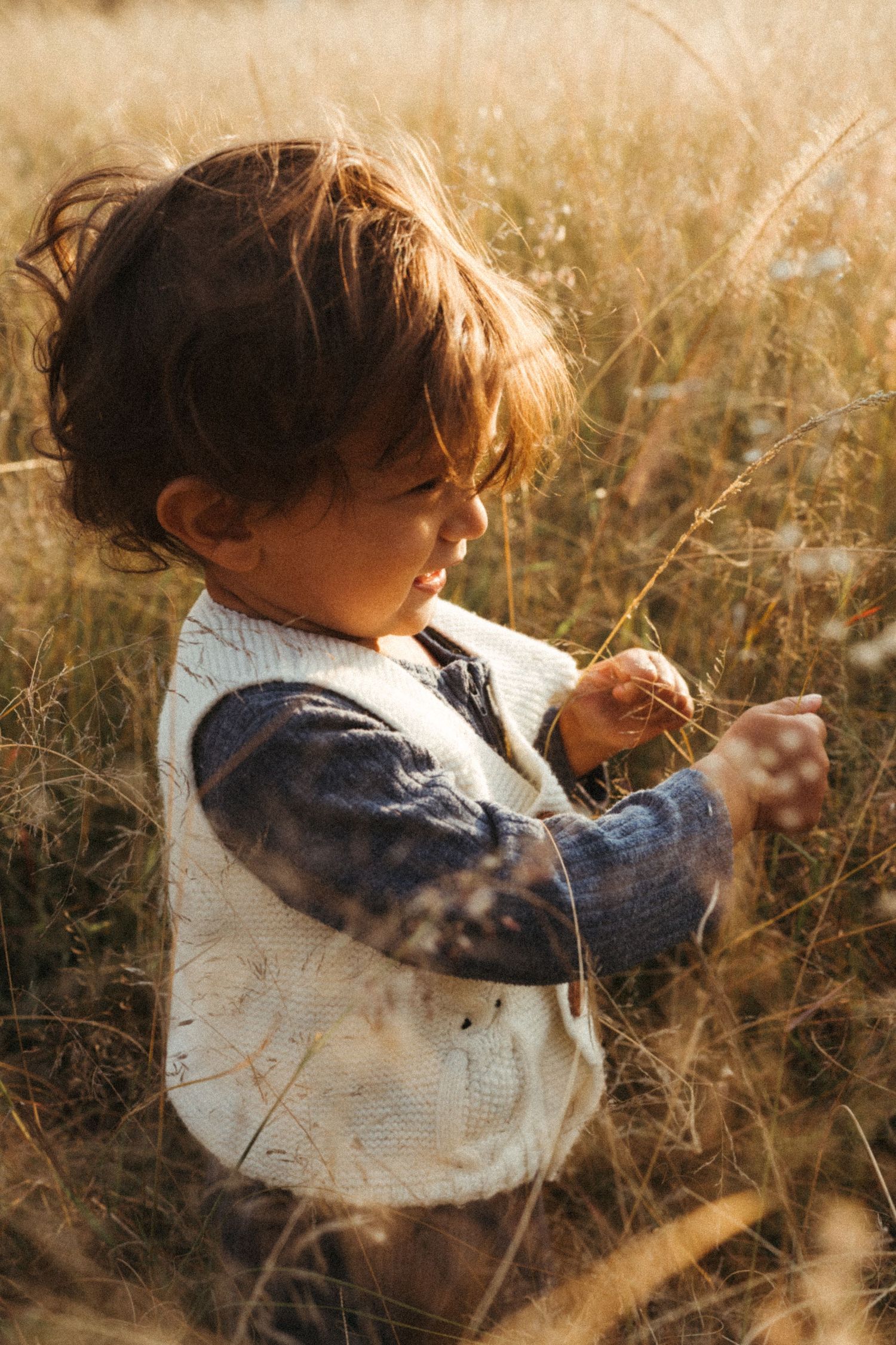 Child in white vest plays among tall grass during sunset.