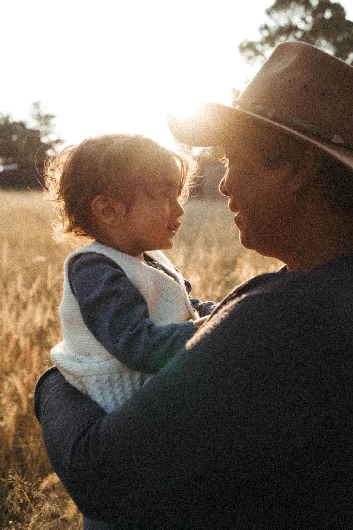 Close-up portrait of two figures in warm sunlight with a brown hat and natural backdrop.