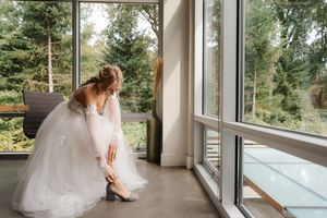 A bride in a white wedding dress sits by large windows overlooking a scenic forest view.