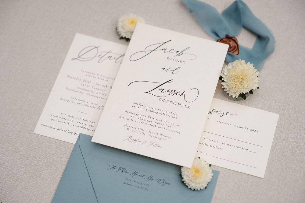 Elegant wedding invitation suite with dusty blue envelopes and white dried flowers on a neutral linen background.