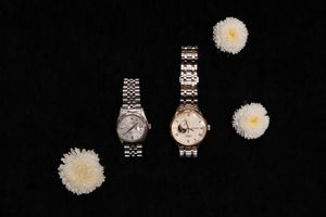 White daisy flowers scattered among silver and gold wristwatches on a dark background create an elegant product display.