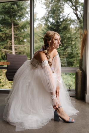 Bride in flowing white tulle gown and pink sweater sits thoughtfully on wooden bench in sunlit room.