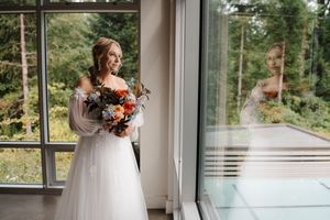 A bride in a white long-sleeved gown holds an autumn-colored bouquet while standing by large windows in natural light.