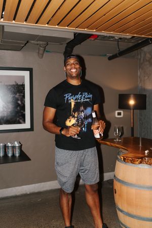Person in black t-shirt and grey shorts smiling while holding wine glasses in a rustic cellar setting.