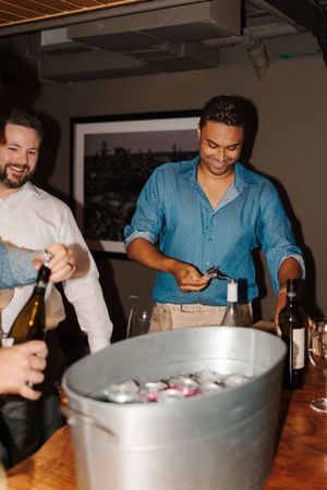 People gather around a large ice bucket at a party table, reaching for drinks and socializing in a dimly lit room.