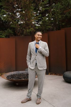 A person in a light gray suit and blue tie stands on a patio near a decorative fire bowl and brown wall.