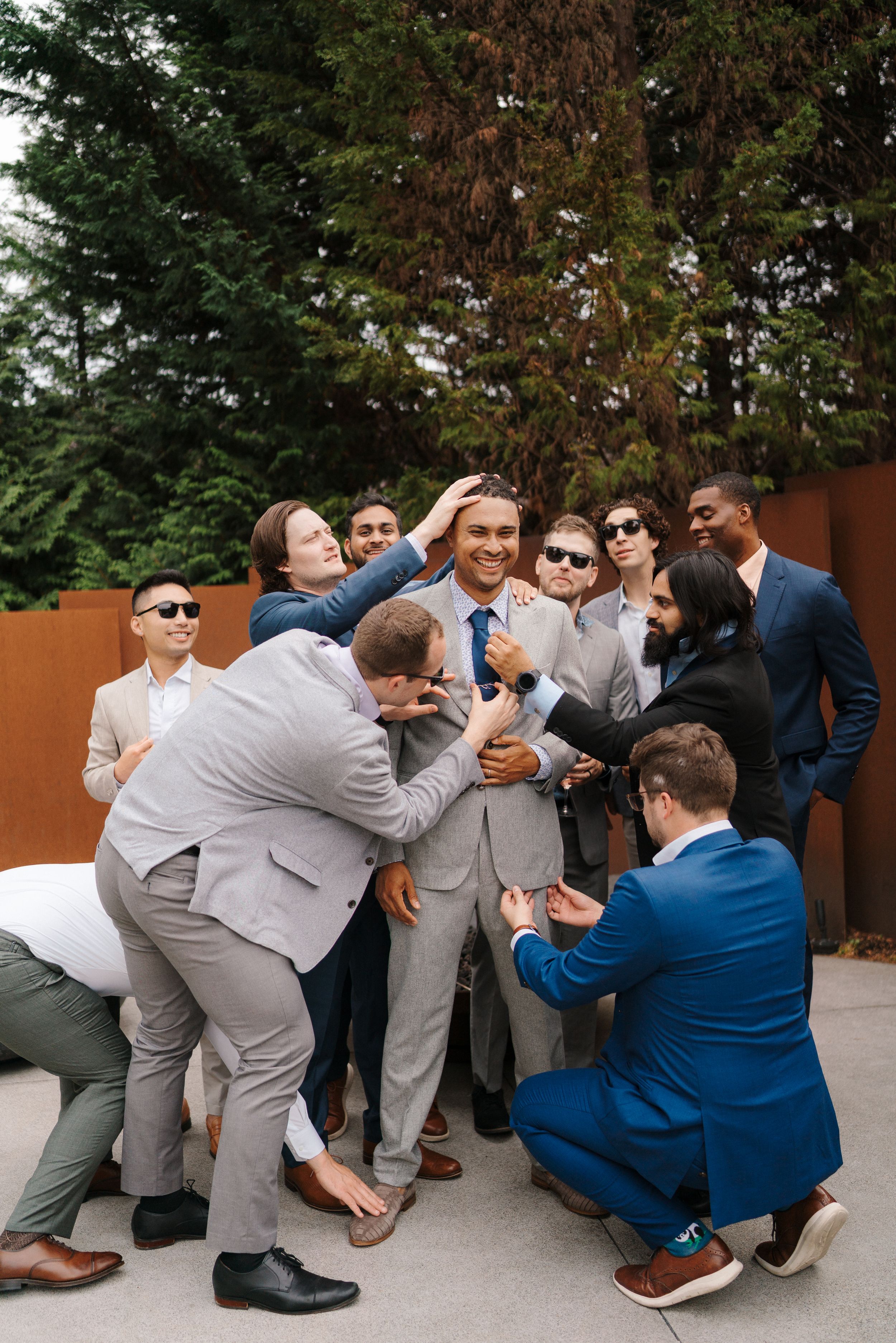 Wedding party members in gray suits and one in blue pose playfully for a series of candid group photos outdoors.