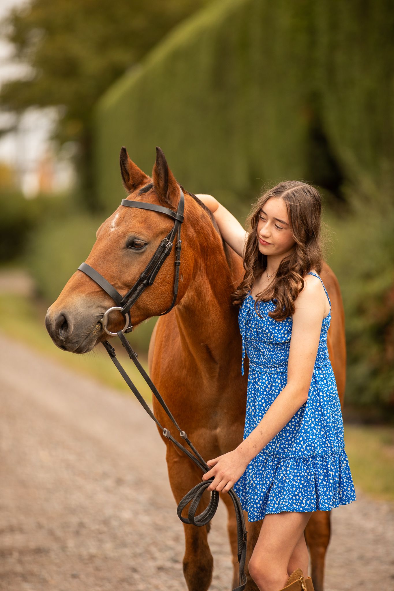 A young rider in a blue polka dot dress poses affectionately with a chestnut brown horse in an outdoor setting.