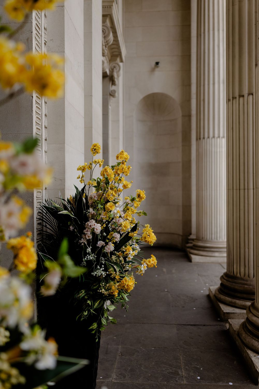 Yellow floral arrangements decorate a classical columned portico with white pillars and stone flooring.