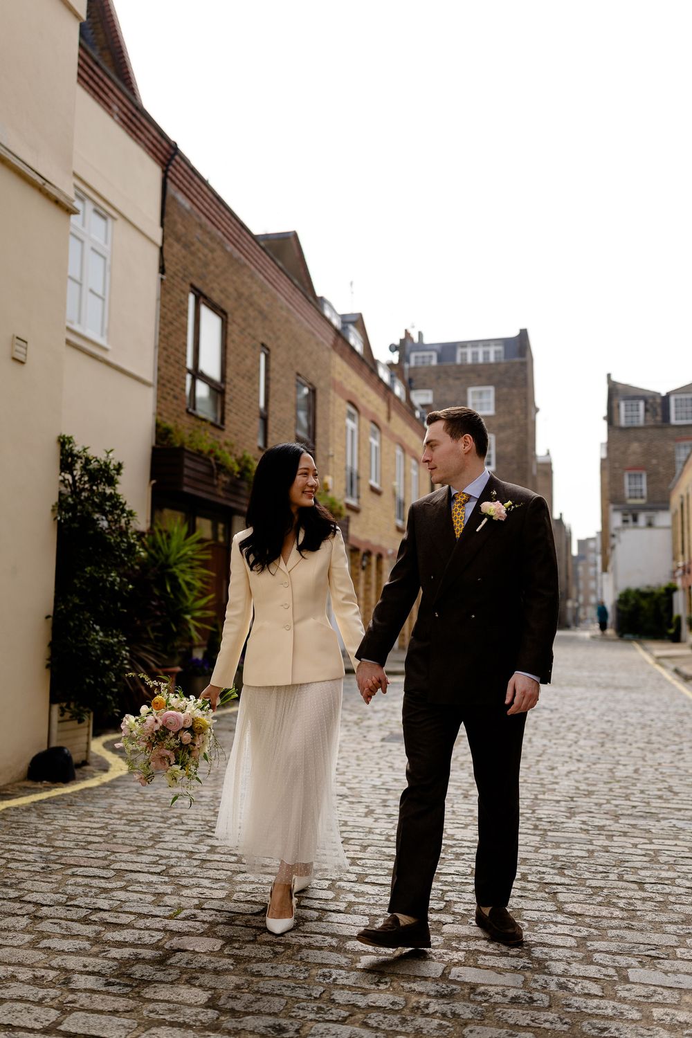 A couple walks hand in hand on a cobblestone street, with the bride wearing a white suit and holding a floral bouquet.