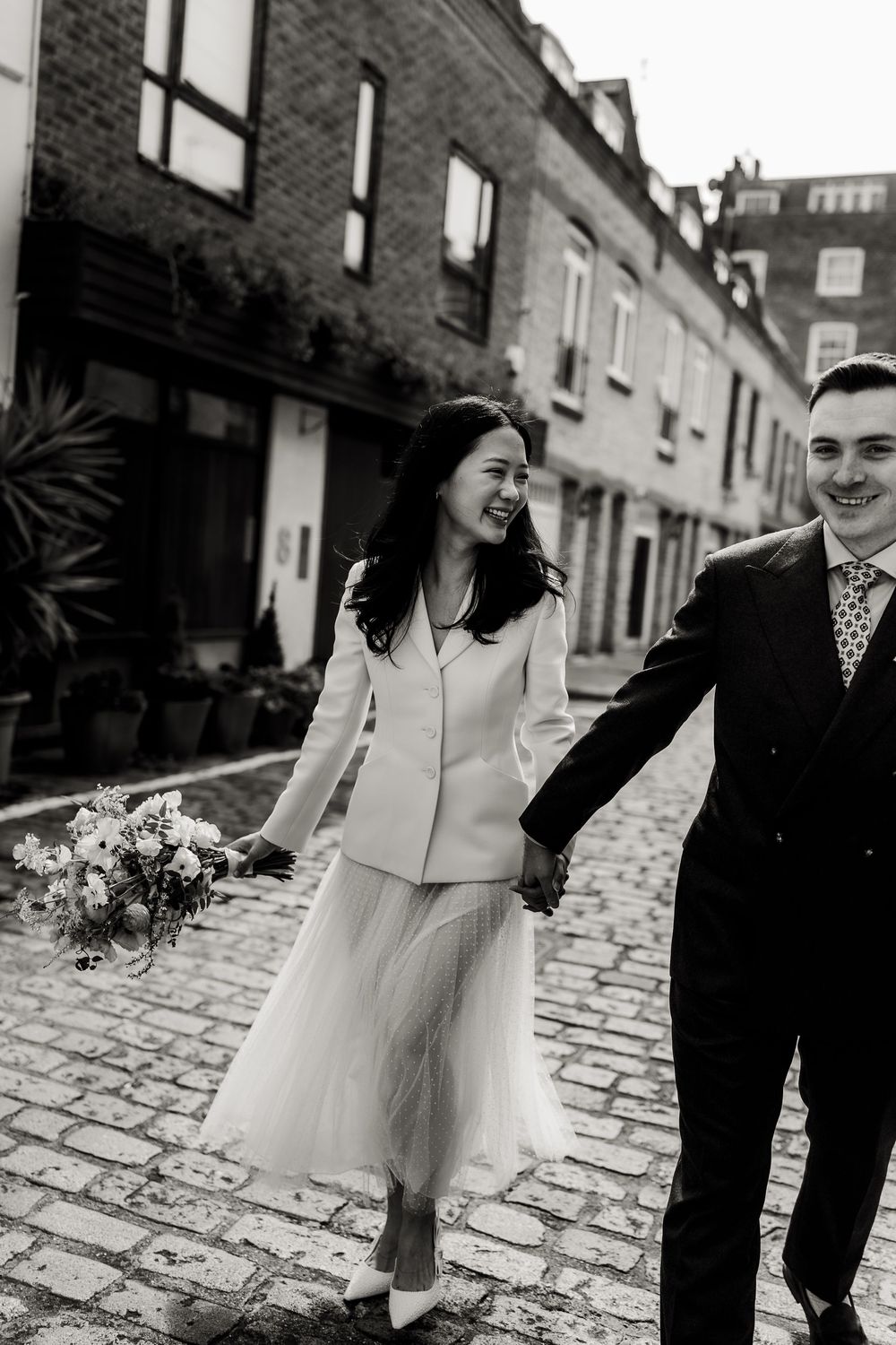 A joyful couple walks down a cobblestone street in wedding attire, with the bride carrying a bouquet of flowers.