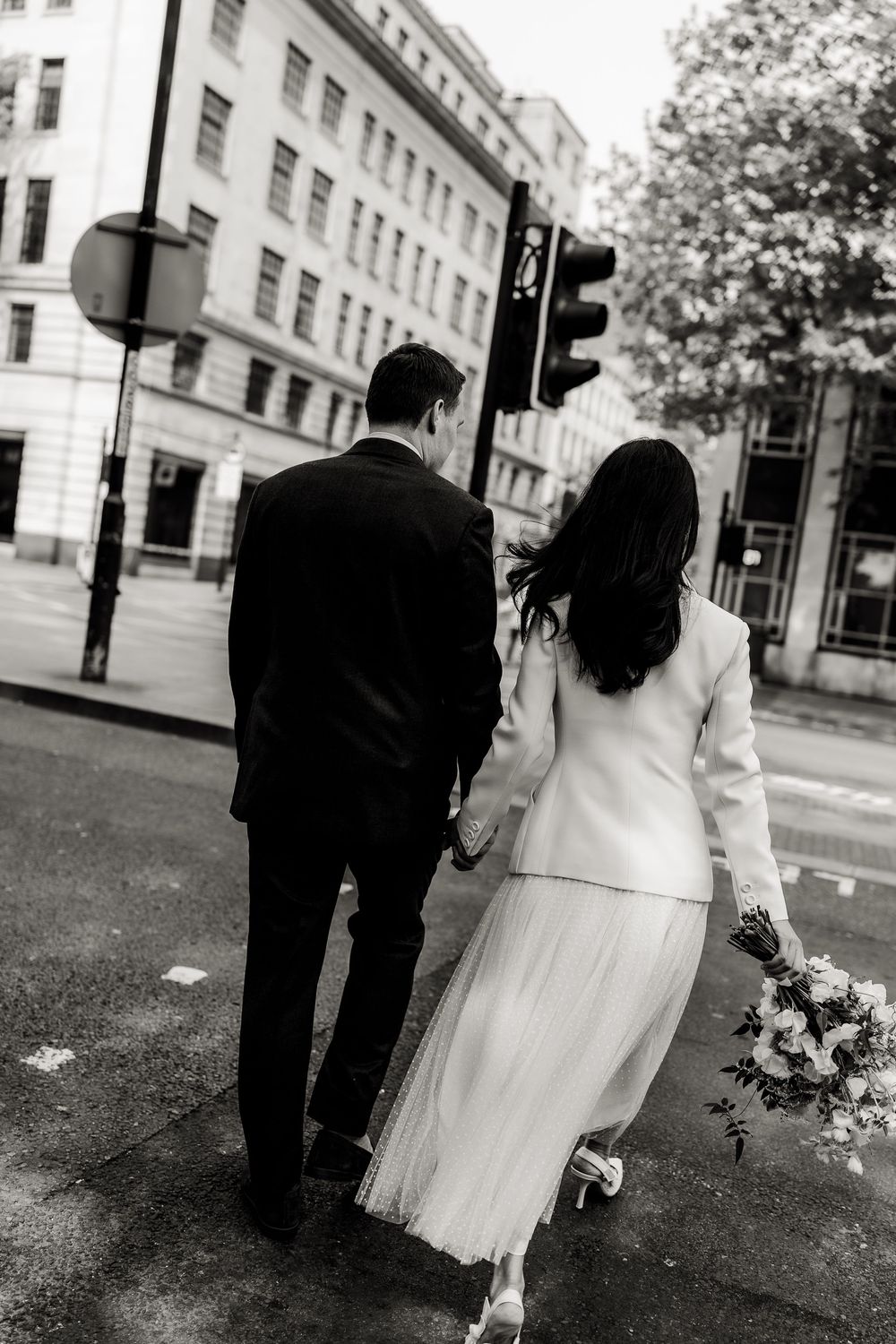 Black and white photo of a couple in wedding attire waiting at a street corner in an urban setting.